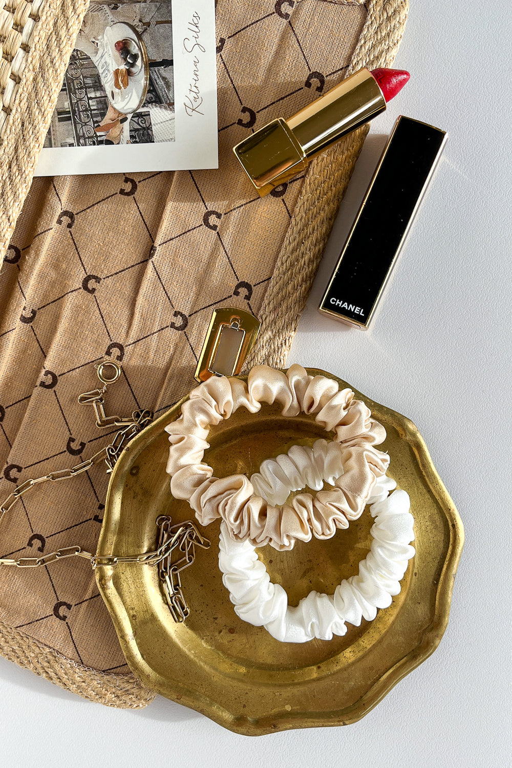 Beige and white natural silk scrunchies on a golden plate next to an open purse and red lipstick