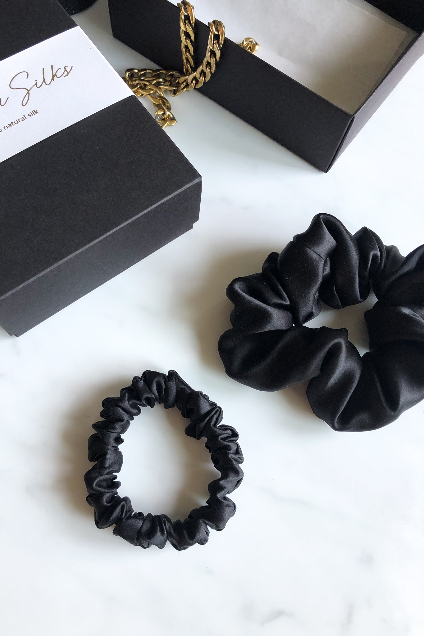 Set of 2 black sillk scrunchies on a marble table, there's Katrina Silks gift box and a golden chain next to it