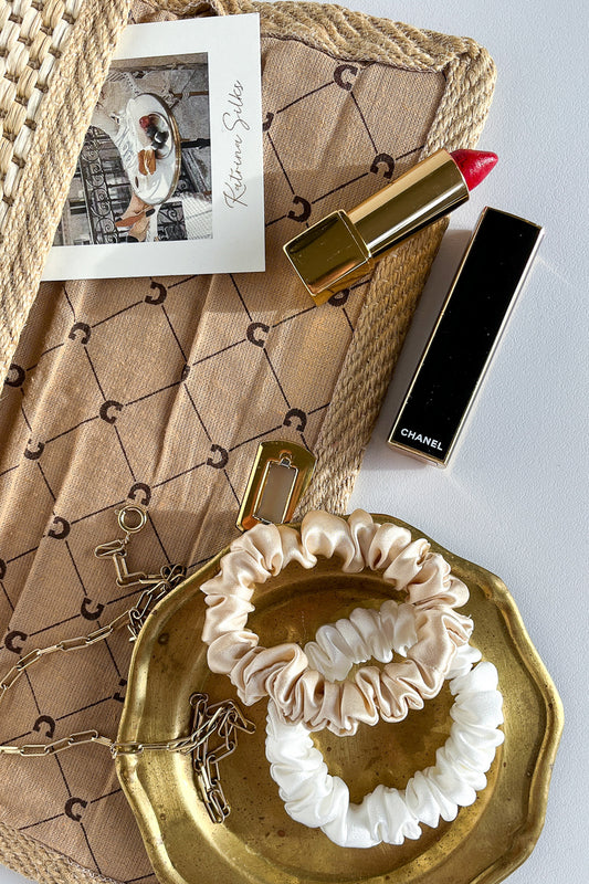 A flatlay look on the table with two silk scrunchies - one beige and one white, red lipstick and a beige, open handbag