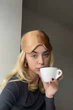 Load image into Gallery viewer, Blond, beautiful girl just awoken with a cup of coffee in her hand and gold colour natural silk eye mask on her forehead.
