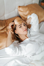 Load image into Gallery viewer, A young woman lying in bed, just waking up, wearing a luxurious silk sleep mask.
