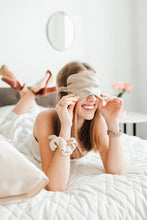 Load image into Gallery viewer, Rising from her sleep, a smiling girl in bed wears a beige natural silk sleep mask.
