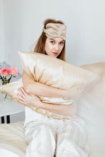 Load image into Gallery viewer, Girl holding beige silk pillowcase and wearing sleep eye mask looking into the camera
