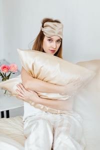 Girl holding beige silk pillowcase and wearing sleep eye mask looking into the camera