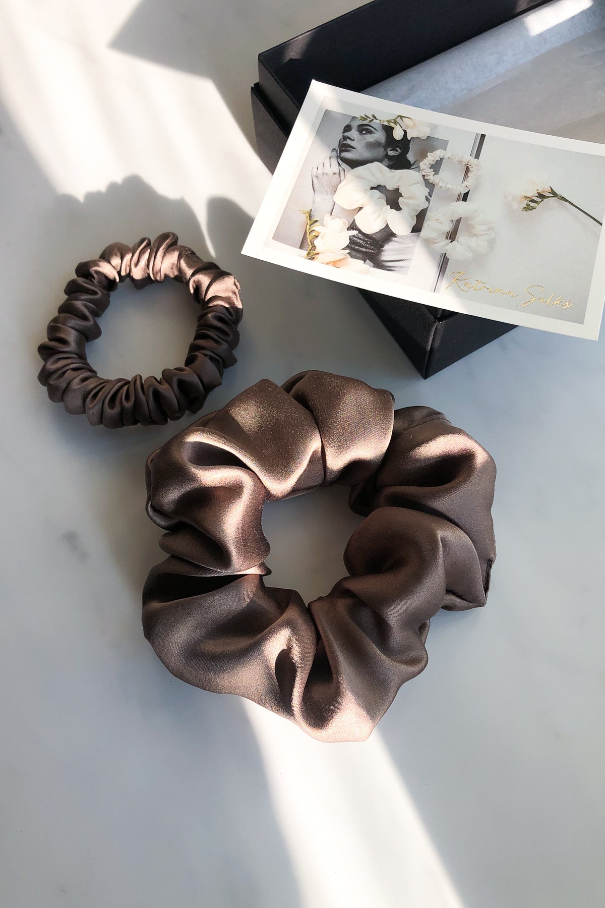 Two mocha colour silk scrunchies in gentle sunlight on a marble table